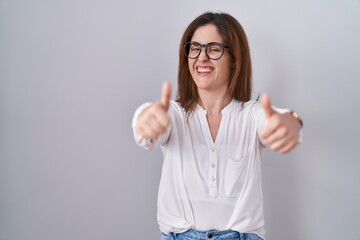 Wall Mural - Brunette woman standing over white isolated background approving doing positive gesture with hand, thumbs up smiling and happy for success. winner gesture.