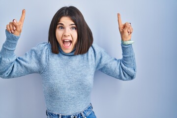 Poster - Young hispanic woman standing over blue background smiling amazed and surprised and pointing up with fingers and raised arms.