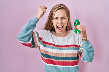 Wall Mural - Young blonde woman holding support green ribbon annoyed and frustrated shouting with anger, yelling crazy with anger and hand raised