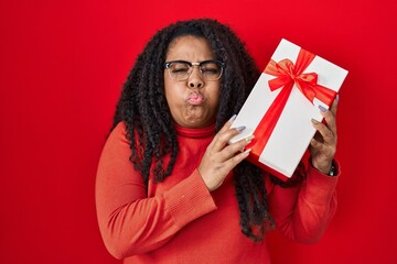 Sticker - Plus size hispanic woman holding gift puffing cheeks with funny face. mouth inflated with air, catching air.