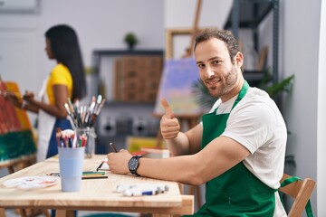 Wall Mural - Hispanic man at painter studio smiling happy and positive, thumb up doing excellent and approval sign