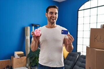 Sticker - Young hispanic man with beard holding piggy bank and credit card at new home celebrating crazy and amazed for success with open eyes screaming excited.