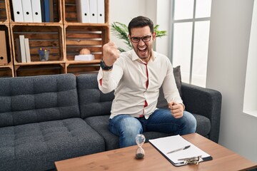 Sticker - Young hispanic man with beard working at consultation office angry and mad raising fist frustrated and furious while shouting with anger. rage and aggressive concept.