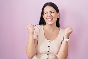 Wall Mural - Young hispanic woman standing over pink background celebrating surprised and amazed for success with arms raised and eyes closed. winner concept.