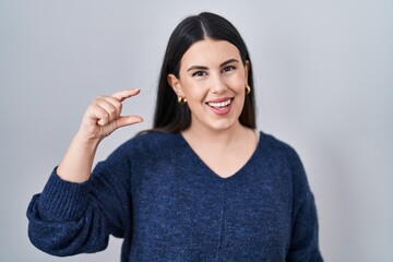 Poster - Young brunette woman standing over isolated background smiling and confident gesturing with hand doing small size sign with fingers looking and the camera. measure concept.