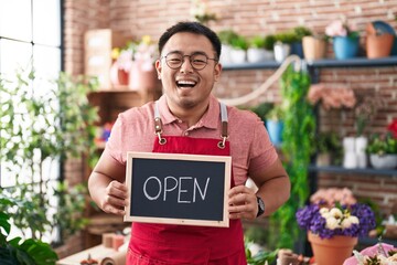 Poster - Chinese young man working at florist holding open sign smiling and laughing hard out loud because funny crazy joke.