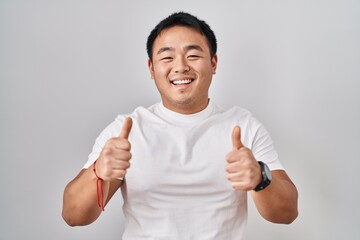 Sticker - Young chinese man standing over white background success sign doing positive gesture with hand, thumbs up smiling and happy. cheerful expression and winner gesture.