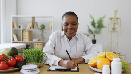 Wall Mural - Portrait of african american woman in lab coat holding pen above clipboard while sitting at writing desk in office. Nutrition professional making changes in patient's diet plan in cozy workplace.