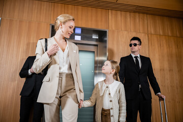 happy mother and child holding hands while standing near hotel elevators and bodyguards in suits, personal protection, successful woman and preteen daughter, family travel, security service