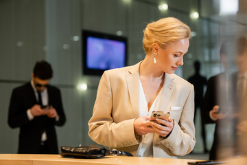 personal security, blonde woman in suit holding smartphone near reception desk, two bodyguards standing on blurred background, digital age, hospitality industry, personal security