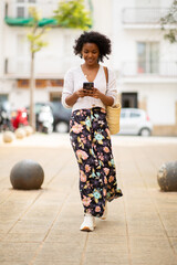 Wall Mural - Full body young african american woman walking in city with mobile phone