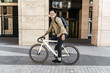A young programmer guy riding to work on a bicycle with a bag, to the office on an eco-transport, a millennial in stylish clothes. Listens to music in wireless headphones playlist from the app.