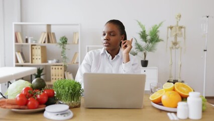 Wall Mural - Attractive multiracial female in white coat working on portable computer in cozy workplace at modern hospital. Mindful dietitian reading about latest researches in nutrition science via internet.