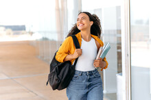 Happy Lovely Curly Haired Brazilian Or Hispanic Female Student, With A Backpack, Hold Books And Notebooks In Her Hand, Walking Near The University Campus, Looks Away And Smile, Finished School Day
