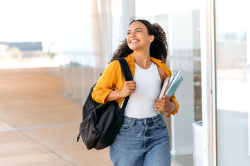 happy lovely curly haired brazilian or hispanic female student, with a backpack, hold books and note