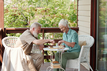 Wall Mural - Happy senior woman with white hair and her husband playing chess while sitting in amchairs by small table on patio of retirement home