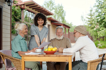 Wall Mural - Young smiling carer in uniform and senior female patient of retirement home looking at paper document next to elderly bearded men