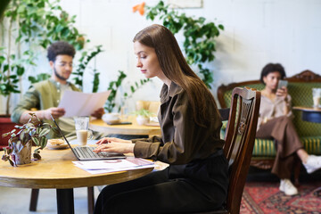 Wall Mural - Young serious female freelancer or solopreneur sitting in cafe in front of laptop, typing an looking at screen while watching online video