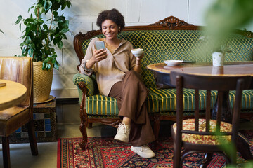 Poster - Happy young restful African American woman scrolling in smartphone or watching online video and having cup of coffee while sitting on couch