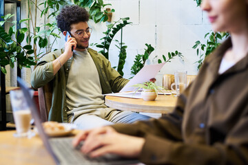 Poster - Young smiling male owner of cafe sitting by table and looking through document while communicating with subordinate on smartphone