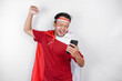 A young Asian man with a happy successful expression while holding his phone and wearing red top, flag headband and cape isolated by white background. Indonesia's independence day concept.
