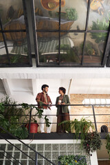 Poster - Below angle of two young employees or friends with coffee having chat while standing by handrail in cafe decorated with green plants