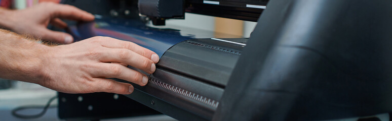 Cropped view of young craftsman holding layer while working with screen printing machine in blurred workshop, small business owner working on project, banner