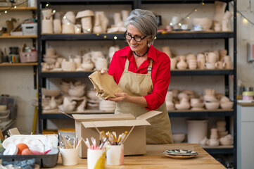 Young seller or worker of earthenware shop packing order of client and wrapping it into paper while standing by table against shelves