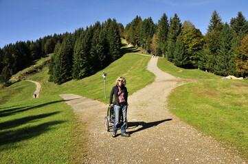 Woman with curls pushing her stroller in the mountains on hiking trails and green meadows with blue sky and forest