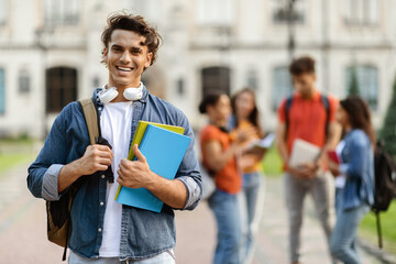 Wall Mural - Handsome Young Male Student With Workbooks In Hands Posing Outdoors At Campus