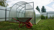 A garden cart near a plastic transparent greenhouse on the green grass on a summer day. Concept gardening and farming