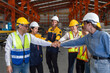 © chadchai - Group of construction workers clad in hard hats and reflective vests, are locked in a cordial fist bump, signaling joint progress. Machinery and steel beams  compose the factory backdrop.