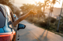 Road Trip, Travel And Vacation Concepts. Happy Woman Hand Out Window Car Blue And Driving On Country Road Into The Sunset. Happy Woman With Sunlight.