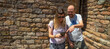 Young happy couple of alternative woman wearing sunglasses and bald man looking laughing and amazed pointing on the mobile phone a route for sightseeing on a sunny day with a romanesque facade behind