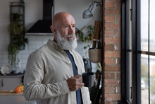 Senior man at home alone looking out of window thinking and having a hot drink