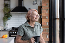 Senior woman at home alone looking out of window happily and having a hot drink