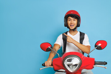 Delivery man in red helmet on red scooter in blue studio, one hand on handle bar another points aside, fast drive concept, copy space