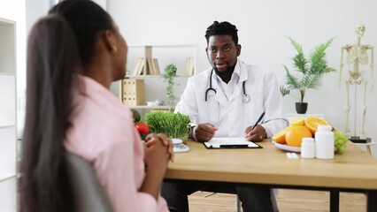 Wall Mural - Confident man with stethoscope writing on paper clipboard during conversation with multiracial woman in office. Serious nutrition expert making notes of patient's medical history for proper meal plan.