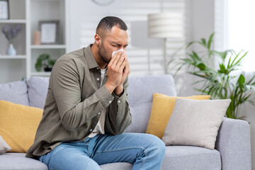 African American man sick at home. He sits on the couch and blows his nose into a tissue