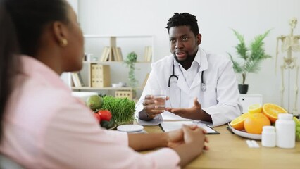 Wall Mural - Attractive bearded man in doctor's coat gesturing at glass with fluid while meeting with multiethnic patient. Smiling nutritionist recommending clean water as help in reducing calorie intake on diet.