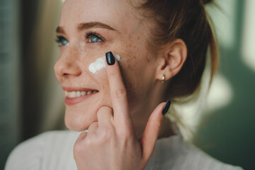 Wall Mural - close-up view of a miling beauty woman applying cream on her freckled cheeks. doing anti aging proce