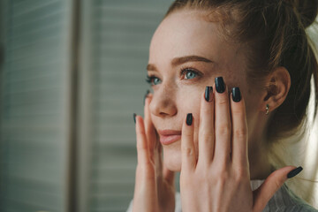 Close-up view of a pretty freckled woman applying facial cream on her face, taking care of her skin sitting in living room at home in the morning. Applying