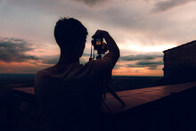 Unrecognizable Man With Camera Taking Picture Of The Evening Orange Sky With Black Clouds