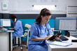Young nurse checking appointments list on digital tablet in busy medical office. Adult woman healthcare specialist working with technology at hospital desk with stethoscope around neck