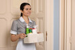 Young chambermaid holding basket with cleaning products in hotel. Space for text