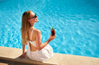 © artiemedvedev - Blonde woman drinks refreshing cocktail sunbathing and sitting near swimming pool at tropical spa. Female in sunglasses enjoys a drink on poolside. Girl chilling in tropical resort on vacation.