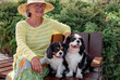 Happy senior woman with hat sitting in a park bench with her two cavalier king charles spaniel dogs. Elderly smiling woman relaxing enjoying retirement