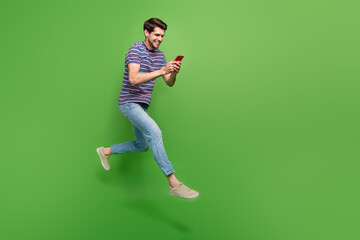 Poster - Full length photo of cute positive man dressed striped t-shirt jumping high texting modern device empty space isolated green color background