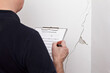 Rental damage concept: man with GERMAN inspection checklist in front of a white wall with a long crack or rip and a piece of plaster missing, .