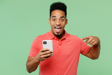 Wall Mural - Young smiling happy fun man of African American ethnicity he wears pink t-shirt hold in hand use point finger on mobile cell phone isolated on plain pastel light green background. Lifestyle concept.